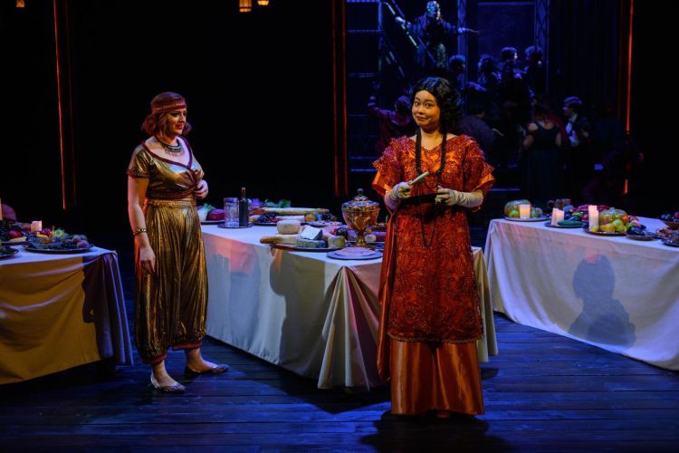 Two opera singers in shiny and embellished costumes with tables of food set for a feast behind them